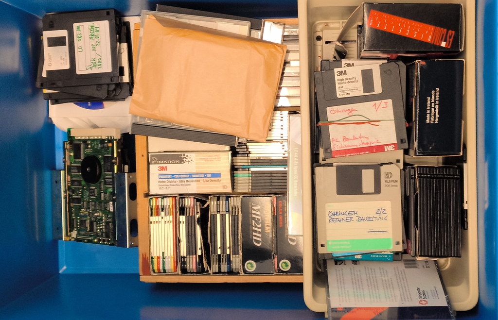 Verschiedene diskette- und CD-ROM-Laufwerke, Speichermedien und Verpackungen in einer blauen Box.