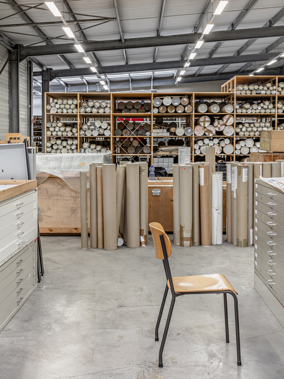 Leeres Büro- oder Lagerzimmer mit Stuhl vor Metallregalen voller Papierrohre und Rollen.